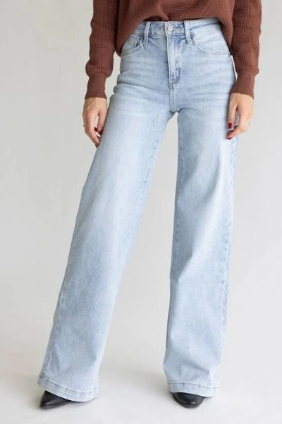 Woman wearing light washed wide leg denim jeans, a long sleeve brown top and pointed black shoes on a plain studio background