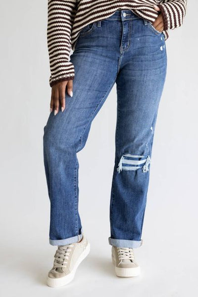 Woman wearing dark wash cuffed straight leg jeans with destressing on the left knew, a striped sweater, and tan sneakers on a plain studio background