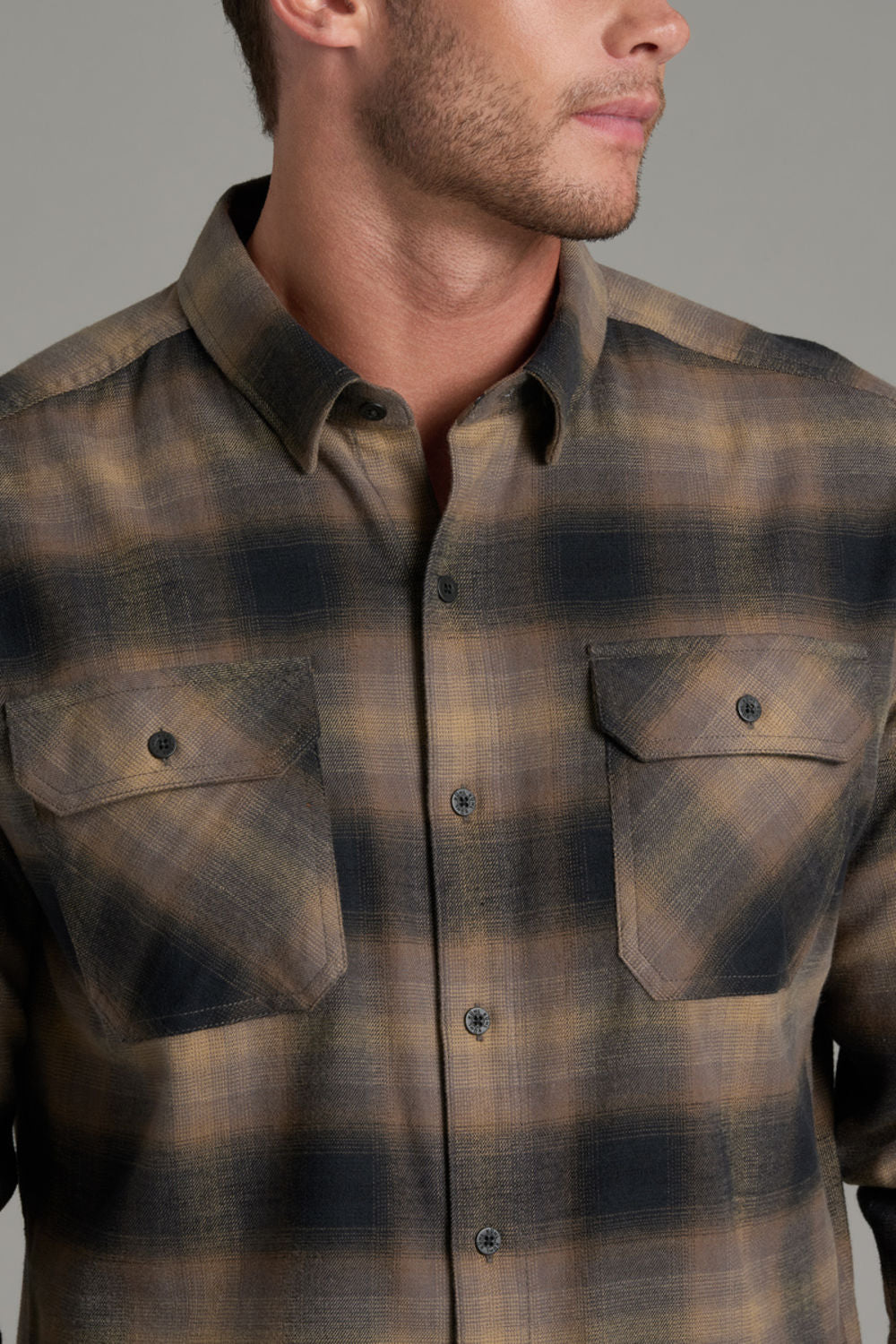 A close-up of a man wearing a brown and dark gray plaid button-down shirt with two chest pockets.