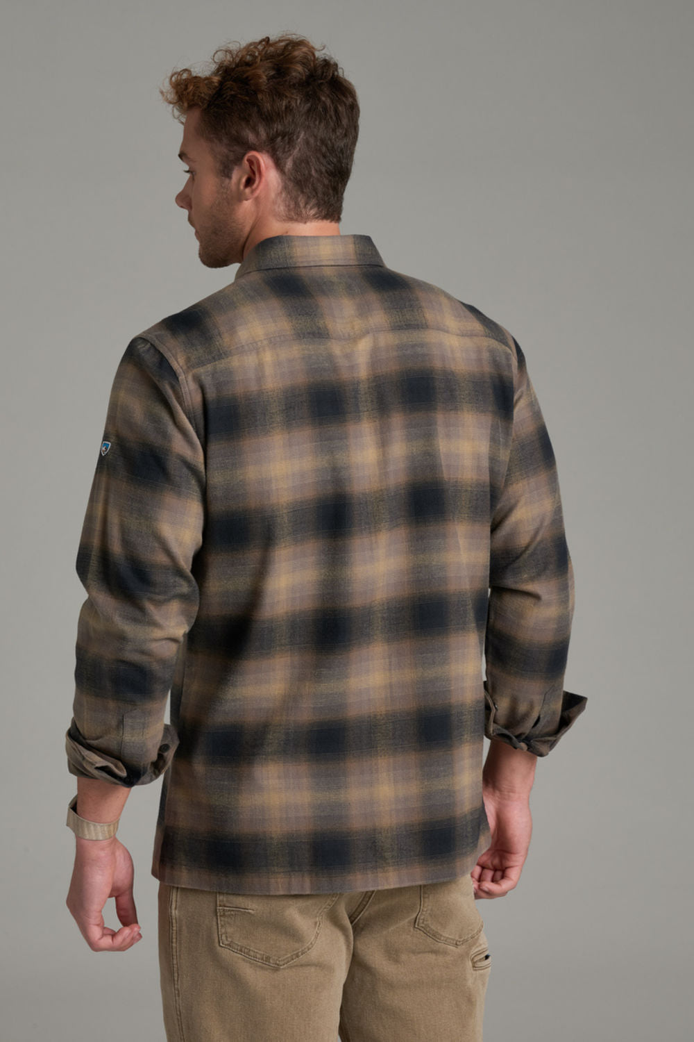 A man viewed from behind wearing a brown and black plaid flannel shirt with tan pants, set against a solid gray studio background.
