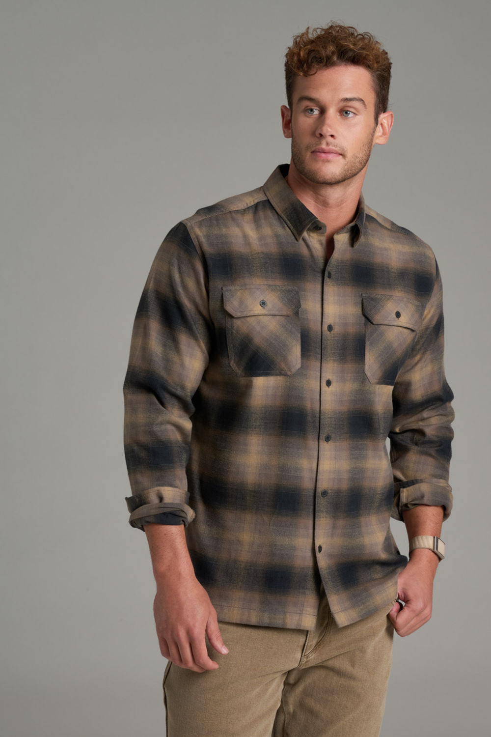 A man wearing a long-sleeve plaid button-up shirt in brown and beige tones, paired with khaki pants, standing against a solid gray studio background.