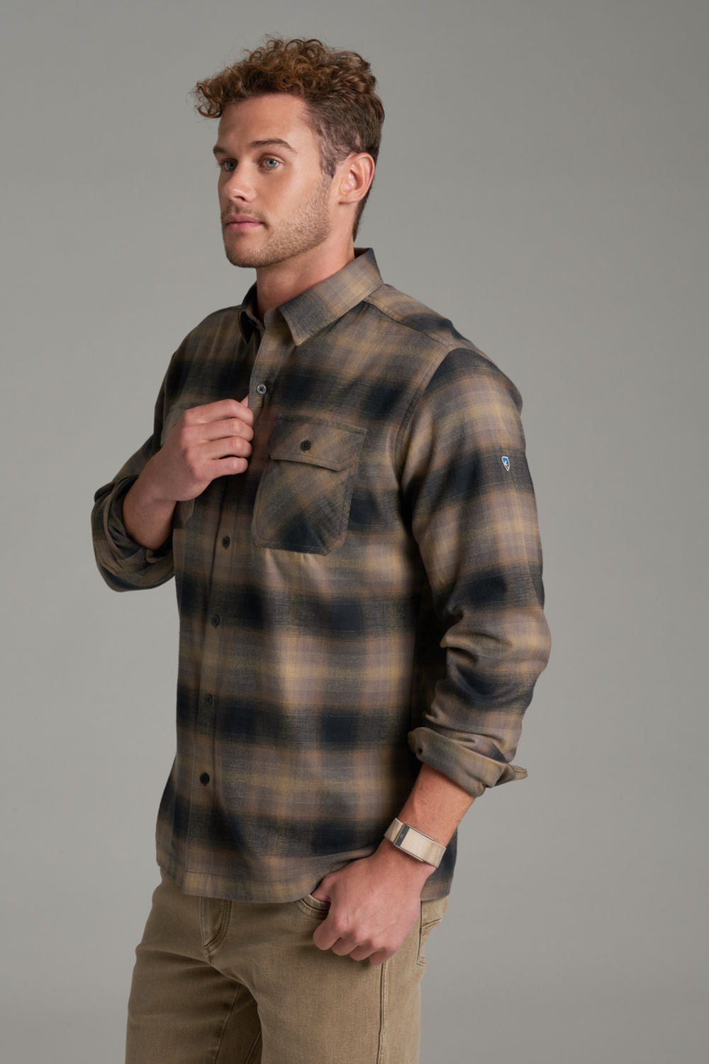 A man wearing a long-sleeve plaid button-up shirt in brown and dark navy tones, paired with khaki pants, photographed against a solid gray studio background.