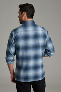 A man wearing a blue and gray plaid button-down shirt with two chest pockets, paired with black pants, standing against a solid gray studio background.