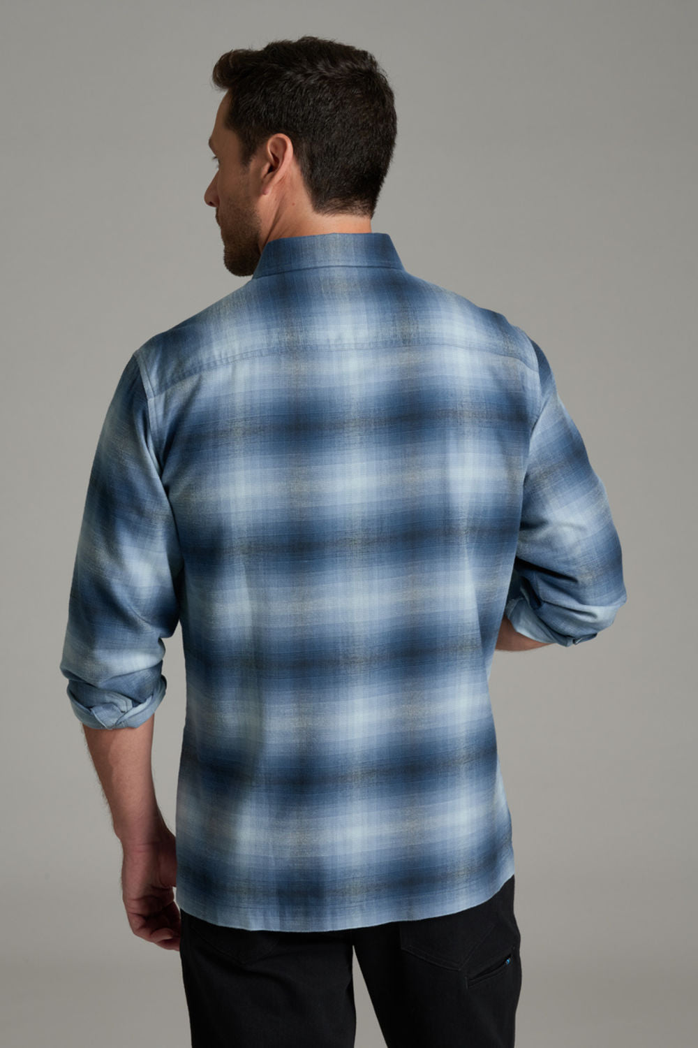 A man wearing a blue and gray plaid button-down shirt with two chest pockets, paired with black pants, standing against a solid gray studio background.