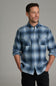 A man wearing a blue and gray plaid button-down shirt with two chest pockets, paired with black pants, standing against a solid gray studio background.