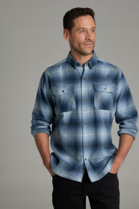 A man wearing a blue and gray plaid button-down shirt with two chest pockets, paired with black pants, standing against a solid gray studio background.
