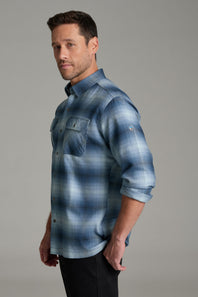A man wearing a blue and gray plaid button-down shirt with two chest pockets, paired with black pants, standing against a solid gray studio background.