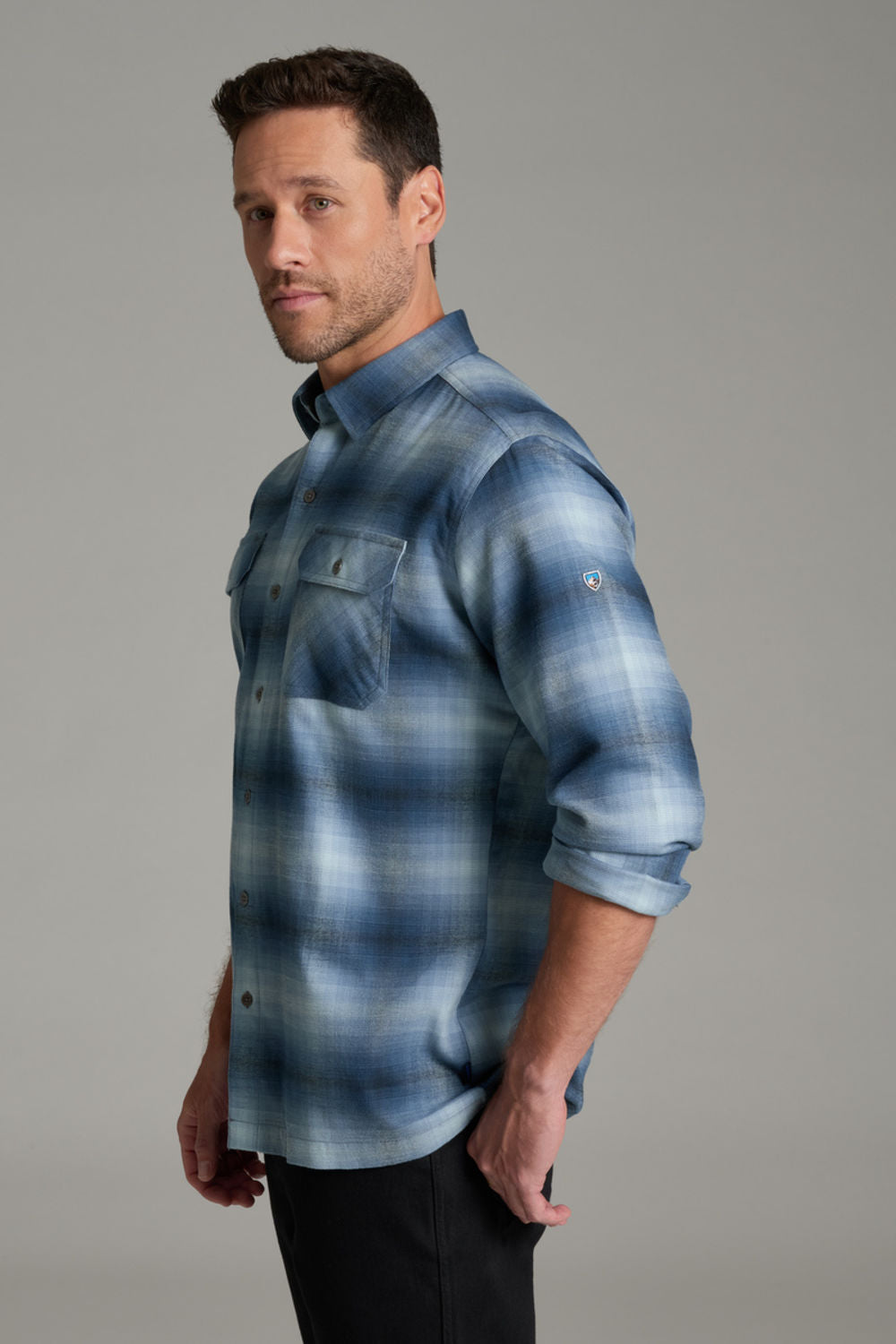 A man wearing a blue and gray plaid button-down shirt with two chest pockets, paired with black pants, standing against a solid gray studio background.