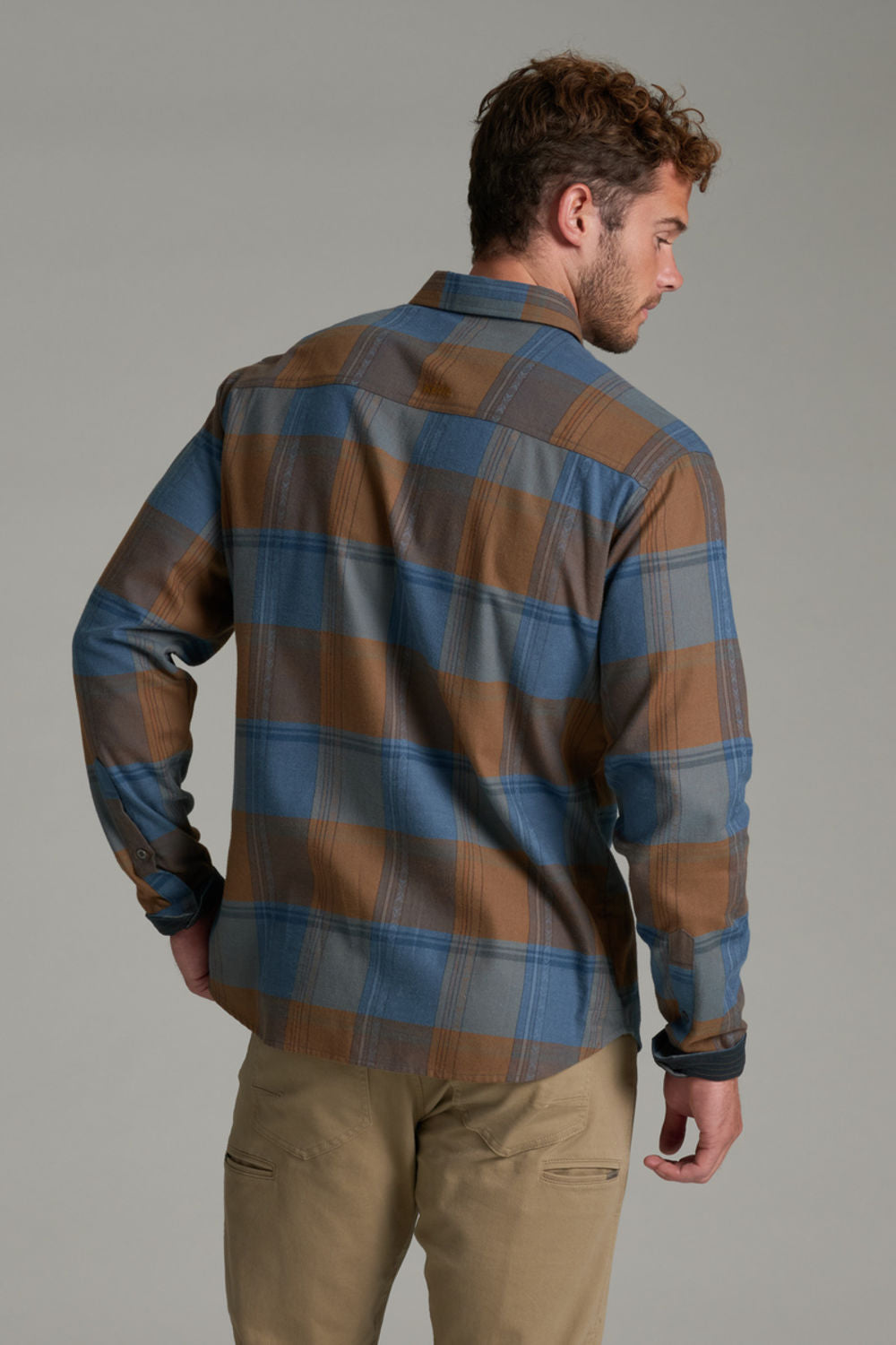 A man wearing a brown and blue plaid button-down shirt, viewed from the back, paired with khaki pants, against a solid gray studio background.