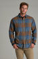 A man wearing a long-sleeve button-up plaid flannel shirt in blue and brown, paired with khaki pants, standing against a solid gray studio background.