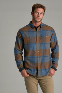 A man wearing a long-sleeve button-up plaid flannel shirt in blue and brown, paired with khaki pants, standing against a solid gray studio background.