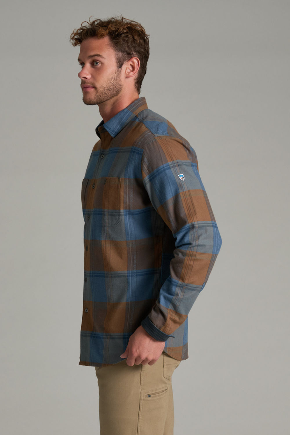 A man wearing a long-sleeve plaid button-down shirt in brown and blue, paired with khaki pants, photographed against a solid gray studio background.
