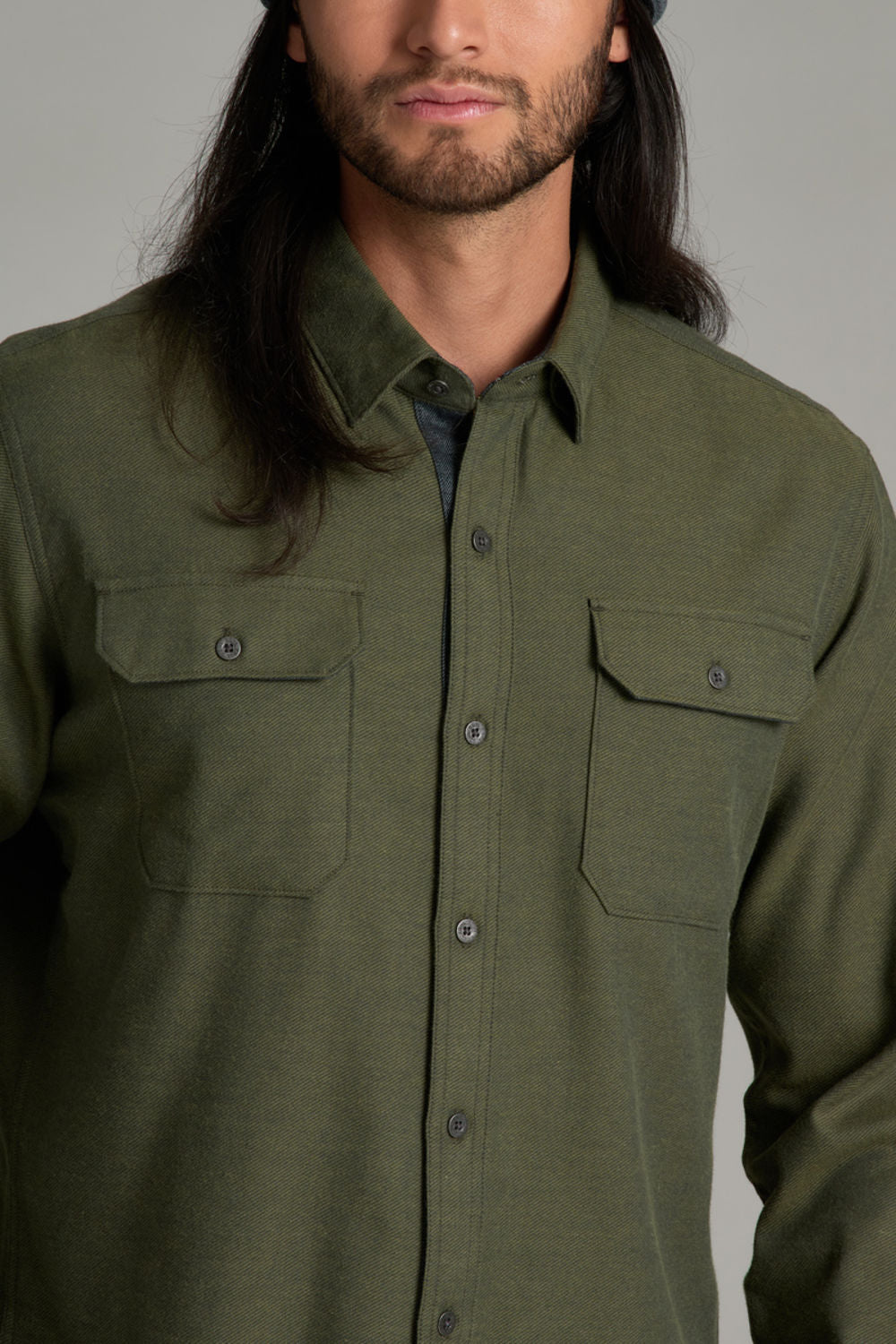 A man wearing an olive green long-sleeve button-up shirt with two chest pockets, shown from the chest up against a plain gray studio background.
