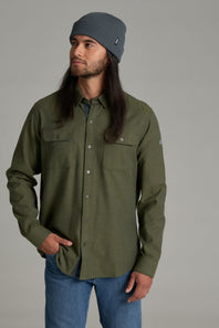 A man wearing an olive green button-down shirt with chest pockets, blue jeans, and a gray beanie, photographed against a solid gray studio background.