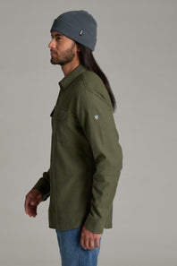 A man in profile wearing an olive green button-up shirt, blue jeans, and a dark gray beanie against a solid gray studio background.