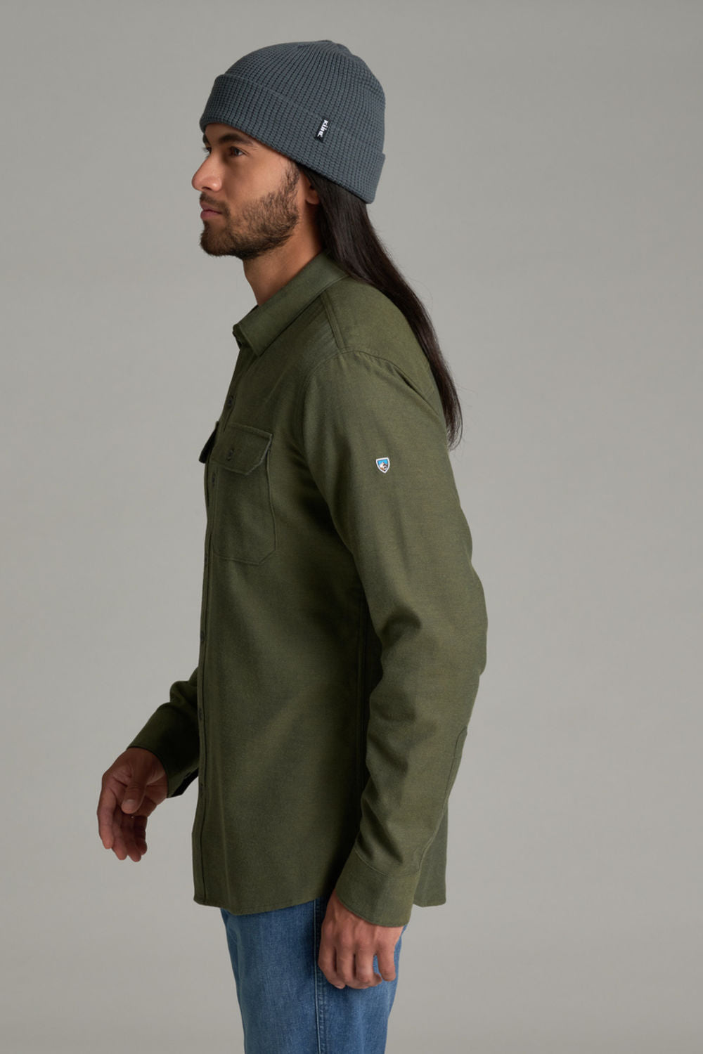 A man in profile wearing an olive green button-up shirt, blue jeans, and a dark gray beanie against a solid gray studio background.