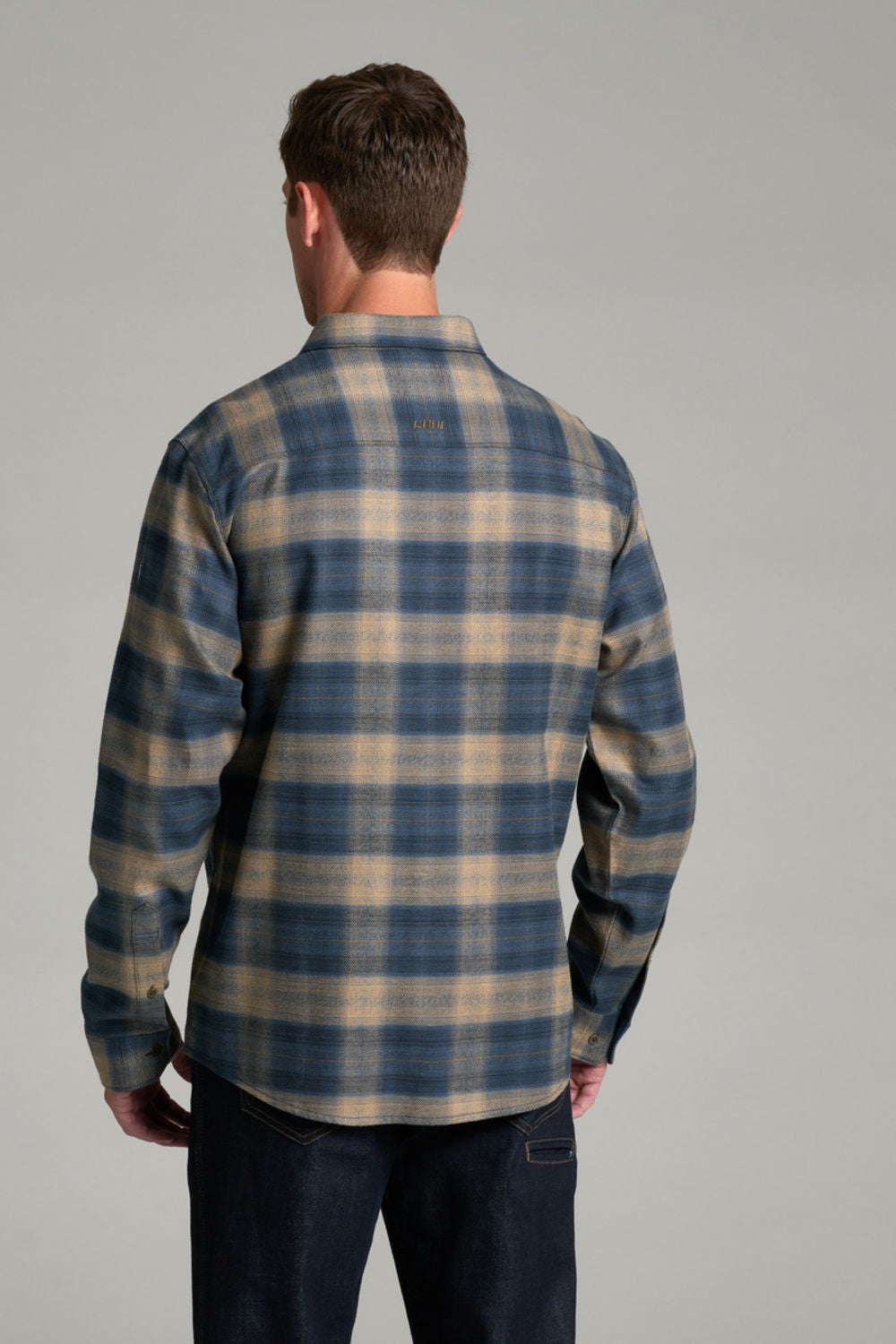A man viewed from behind wearing a blue and beige plaid flannel shirt and dark jeans, standing against a neutral gray studio background.