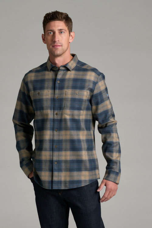 A man wearing a long-sleeve plaid button-up shirt in blue and tan, paired with dark jeans, standing against a solid gray studio background.