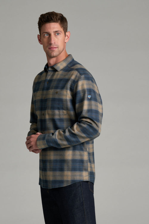 A man wearing a long-sleeve plaid button-up shirt in navy blue and tan, paired with dark jeans, standing against a solid gray studio background.