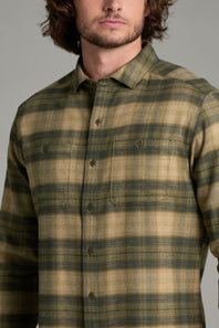 A man wearing a long-sleeve button-up flannel shirt with a green and tan plaid pattern, shown from the chest up against a neutral gray background.