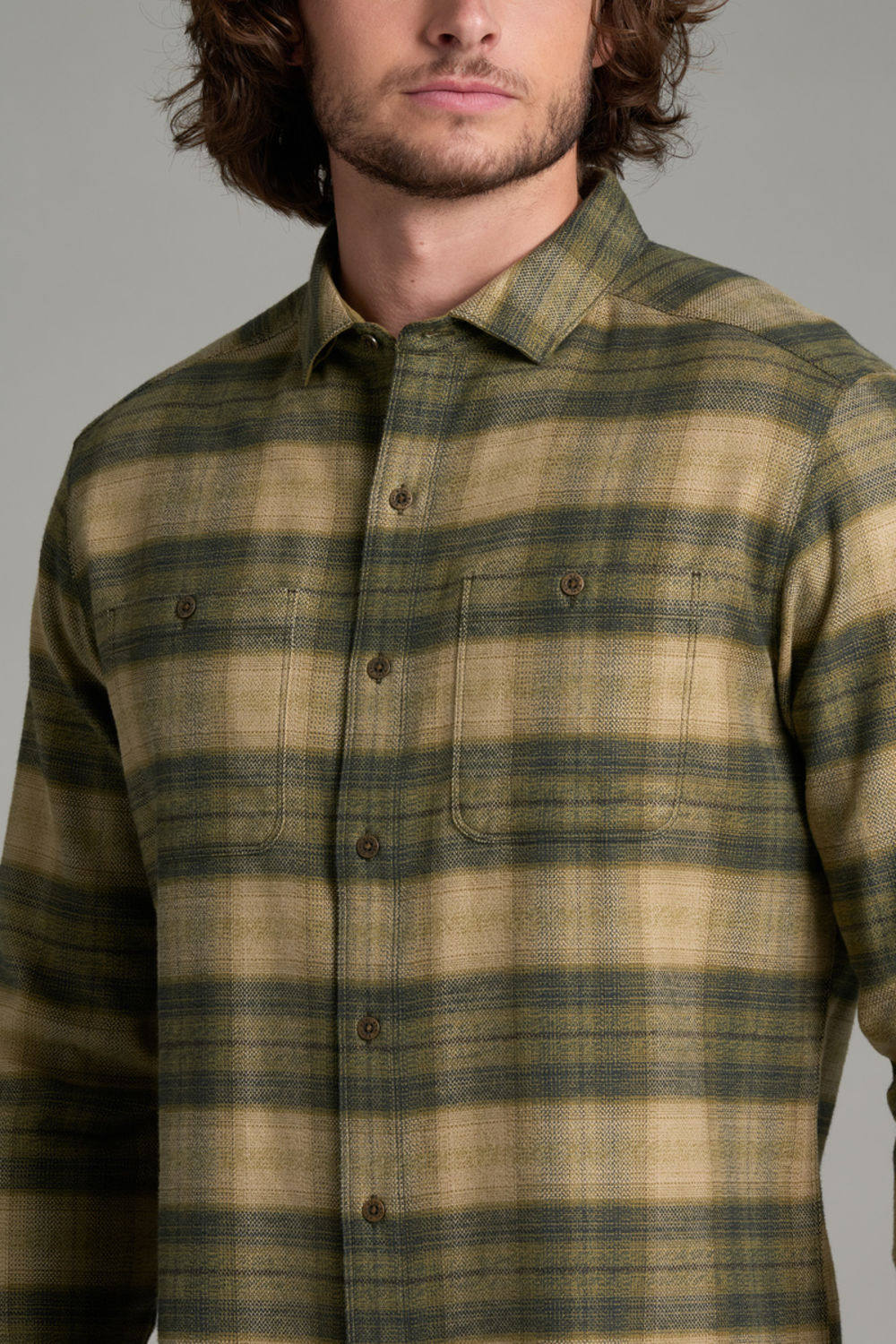A man wearing a long-sleeve button-up flannel shirt with a green and tan plaid pattern, shown from the chest up against a neutral gray background.