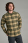 A man wearing a long-sleeve button-up flannel shirt with a green and tan plaid pattern, paired with dark gray pants, photographed against a solid gray studio background.