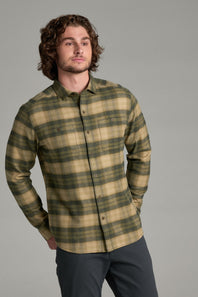 A man wearing a long-sleeve button-up flannel shirt with a green and tan plaid pattern, paired with dark gray pants, photographed against a solid gray studio background.