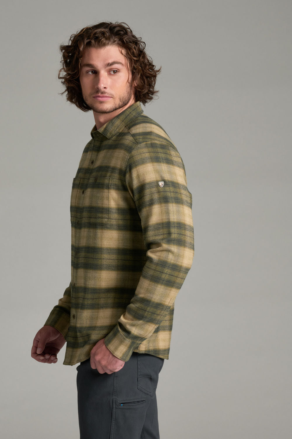 A man wearing a long-sleeve plaid button-up shirt in olive green and tan, paired with dark gray pants, photographed against a solid gray studio background.