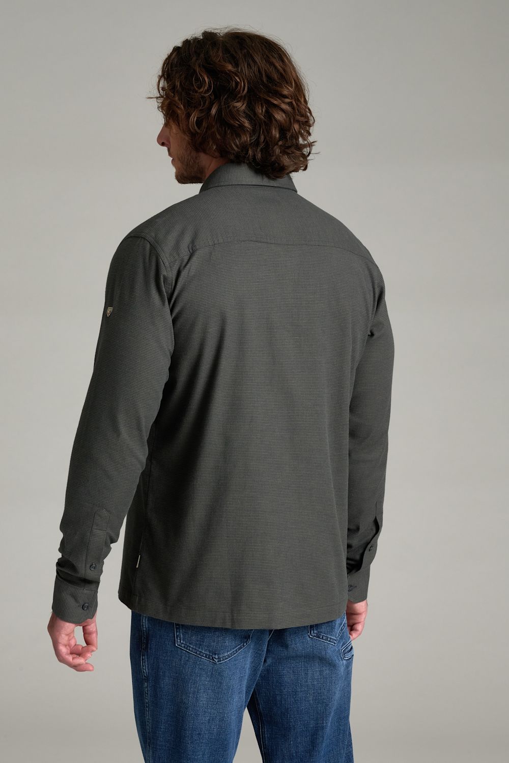 A man with curly brown hair is shown from the back wearing a dark gray long-sleeve button-up shirt and blue jeans against a neutral gray background.