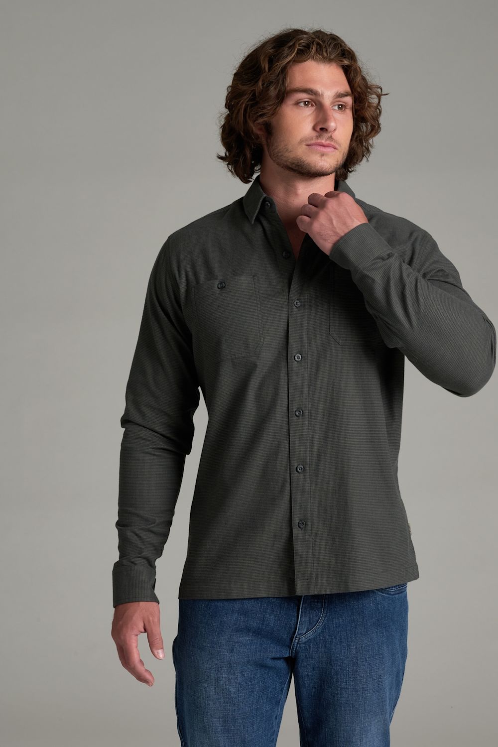 A man wearing a dark charcoal gray long-sleeve button-down shirt and blue jeans, standing against a solid gray studio background.