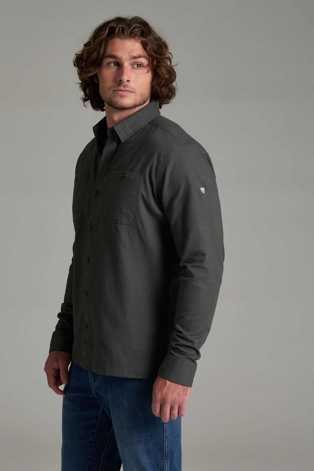 A man wearing a dark charcoal gray long-sleeve button-down shirt with blue jeans, standing against a solid gray studio background.