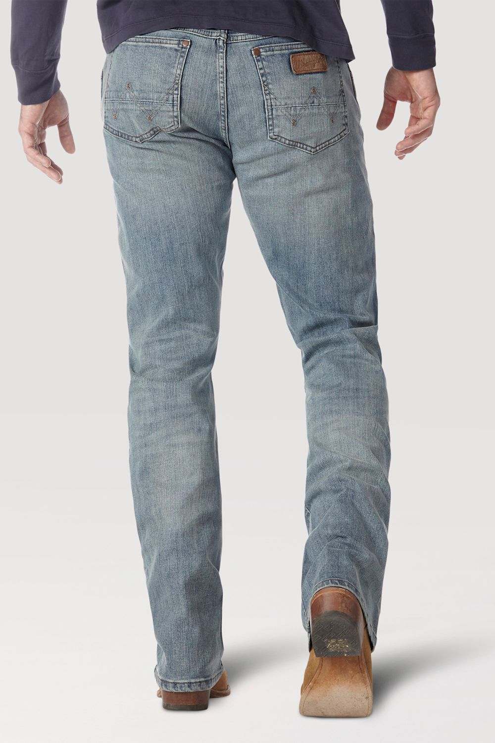 Rear view of a person wearing light-wash blue denim jeans with brown leather boots and a dark navy long-sleeve top, shown against a light gray studio background.