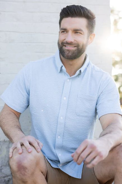 Man wearing a blue button up shirt and khaki shorts sitting in front of a white brick wall