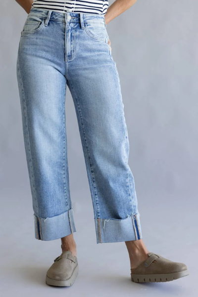 Front view of a model wearing light wash high-rise straight-leg jeans with wide cuffed hems featuring released-edge detailing, paired with taupe slip-on clogs and a striped top against a neutral gray background.