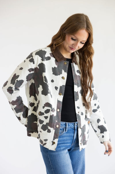 Woman standing in front of a plain studio background wearing a white and brown cow print jacket with a black undershirt and medium was jeans