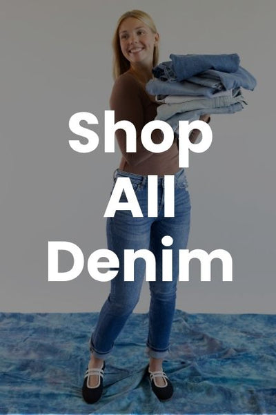 A smiling woman wearing a brown long-sleeve top, blue jeans, and black flats holding a stack of folded denim jeans. Large white text reading Shop All Denim is centered over the image.