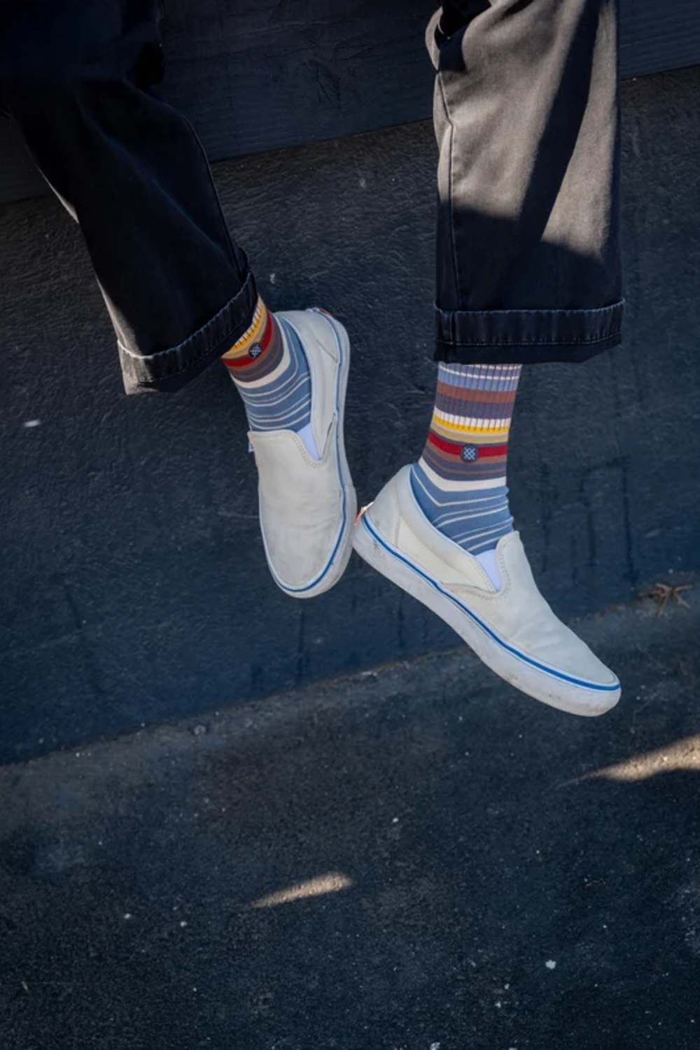 A person's legs dangling in the air, wearing white slip-on canvas sneakers and striped crew socks with a small red heart detail.