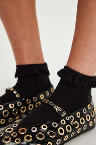 Black loafers with gold eyelets and ruffled black socks on a neutral background