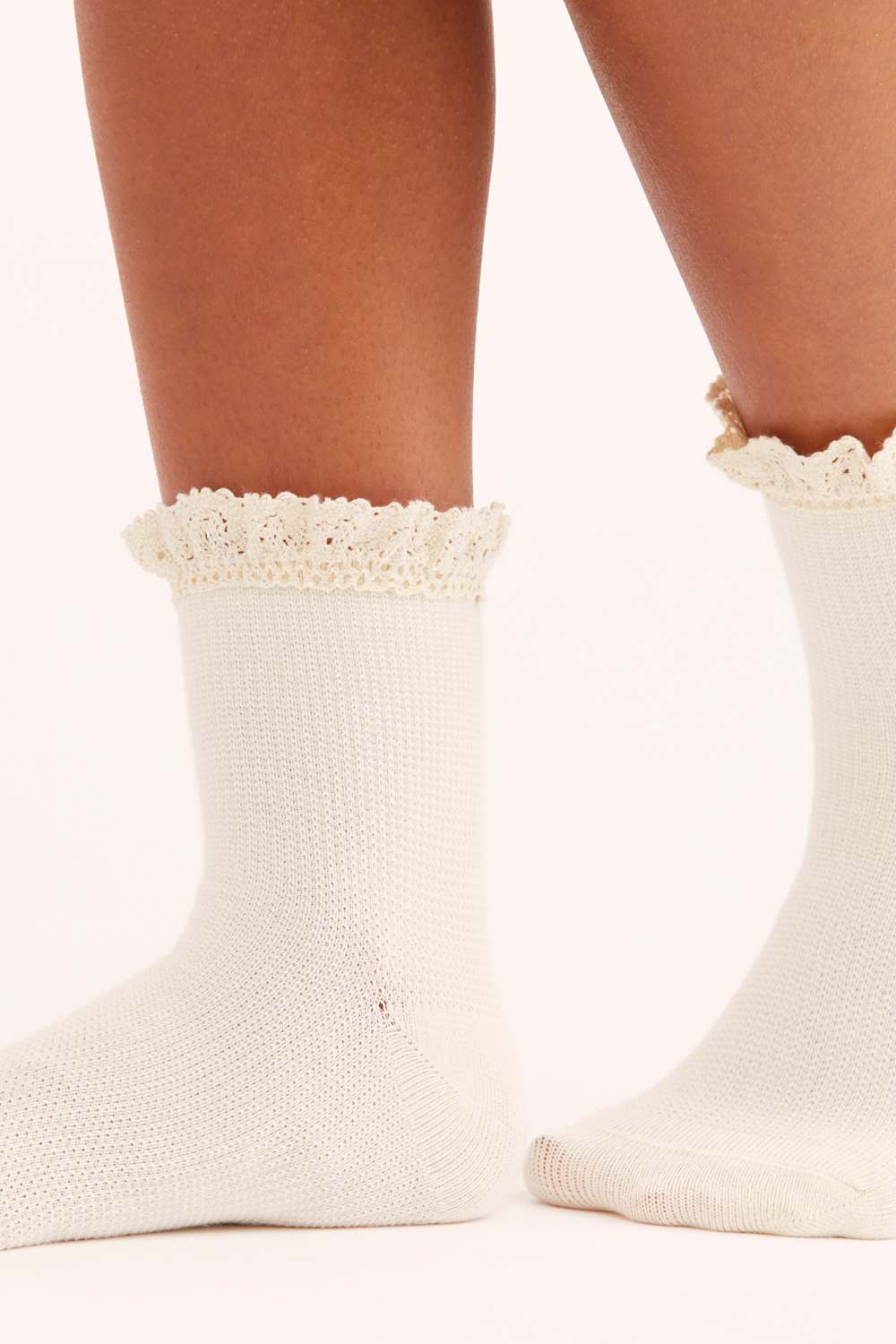 A close-up of a person's feet wearing off-white knit ankle socks with delicate floral lace ruffles at the top, set against a plain white background.