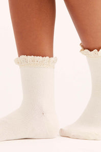 A close-up of a person's feet wearing off-white knit ankle socks with delicate floral lace ruffles at the top, set against a plain white background.
