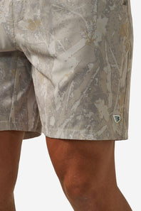 Close-up of a person wearing camouflage athletic shorts in a light beige, gray, and brown pattern, with a small shield logo patch on the lower left leg.