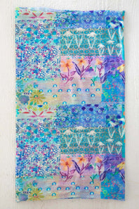 A square fabric art piece featuring a colorful patchwork design with floral and geometric patterns in shades of blue, purple, teal, and pink, displayed flat against a white textured background.