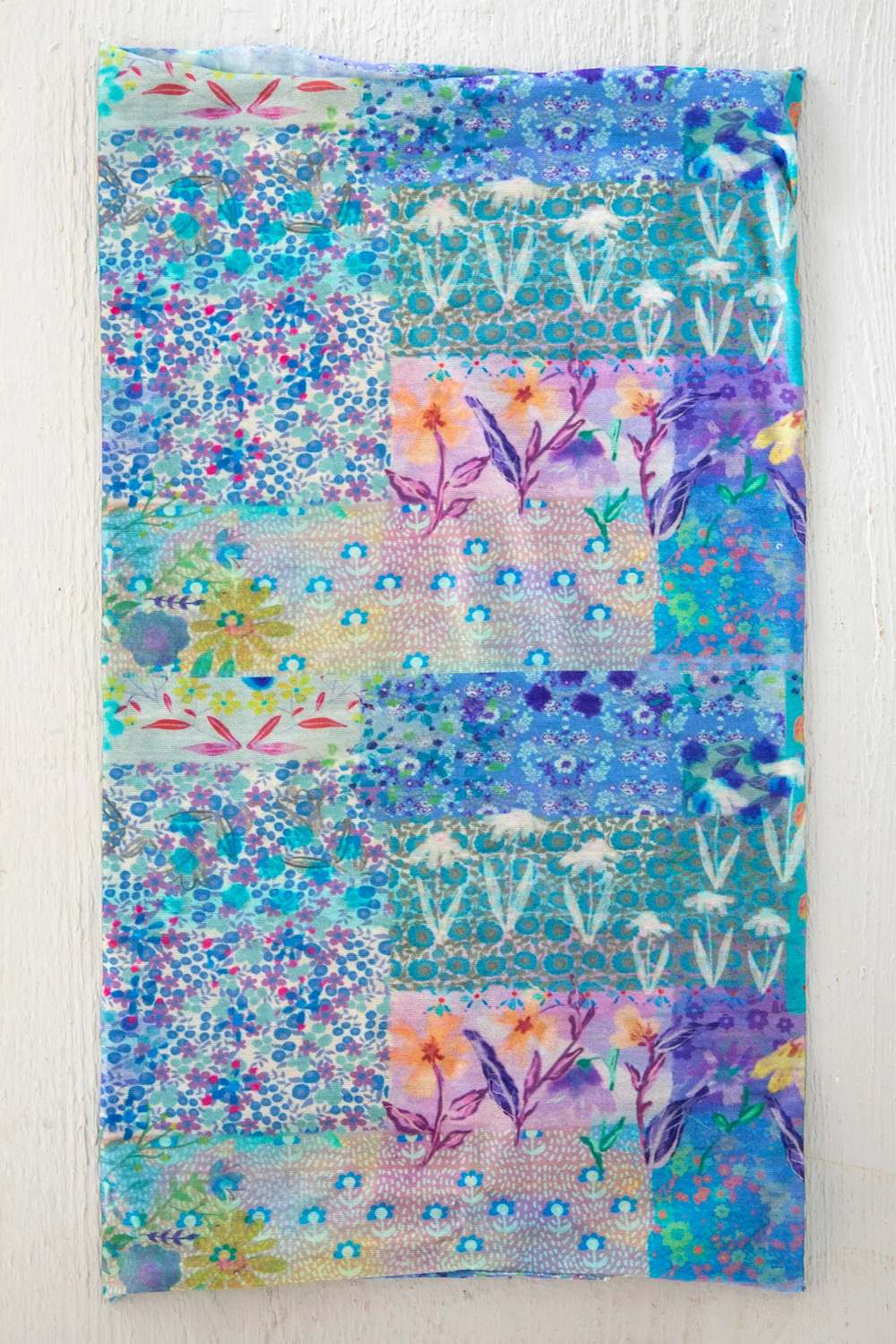 A square fabric art piece featuring a colorful patchwork design with floral and geometric patterns in shades of blue, purple, teal, and pink, displayed flat against a white textured background.