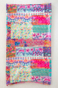 A square patchwork fabric napkin featuring a vibrant multicolor floral and geometric print, displayed flat against a light wood background.