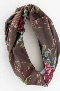 A brown infinity scarf with a colorful floral and embroidered-style pattern, displayed on a white mannequin torso against a white wall.