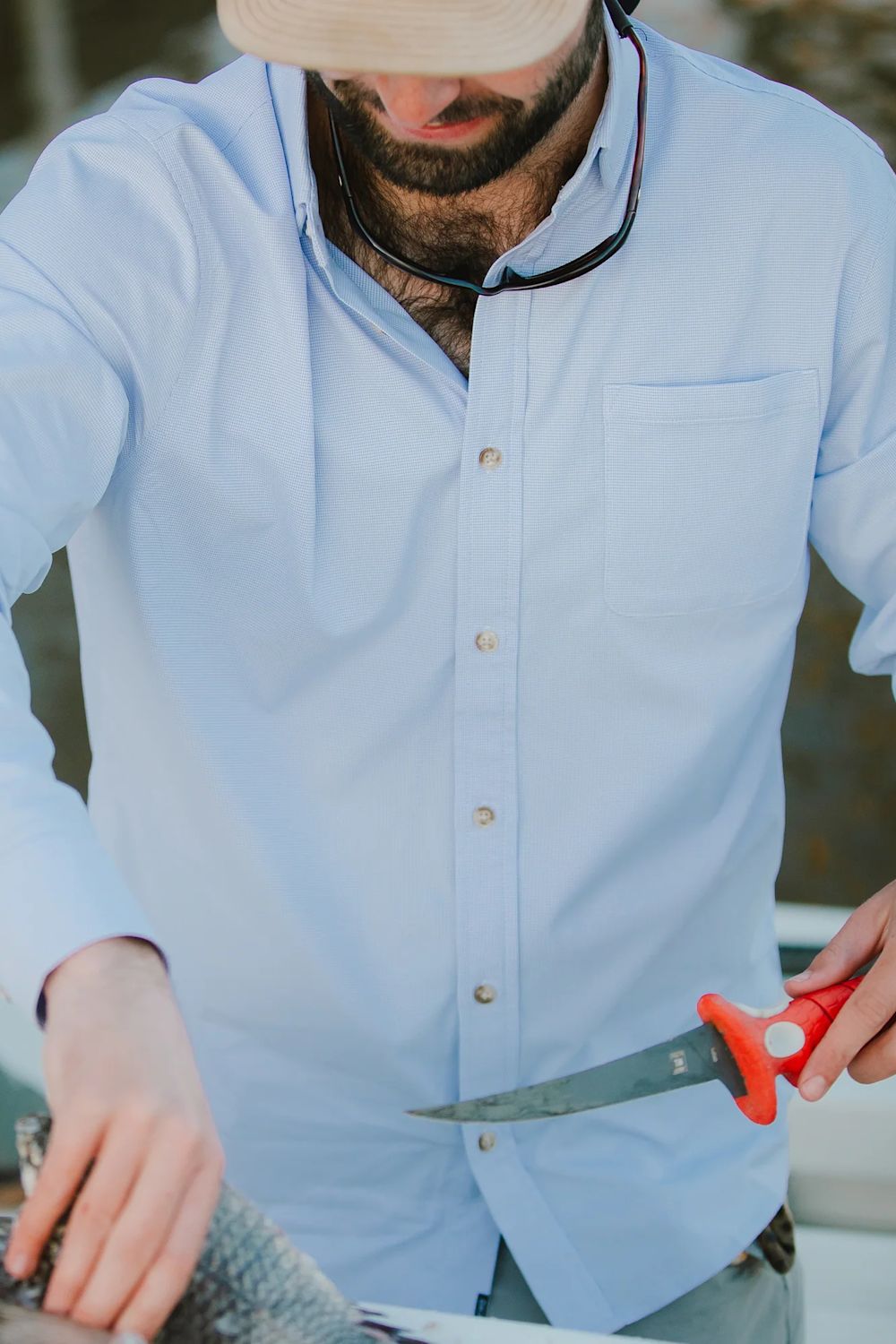 A man wearing a light blue button-down shirt and a beige hat is shown from the chest down, holding a red-handled knife while handling a fish outdoors.