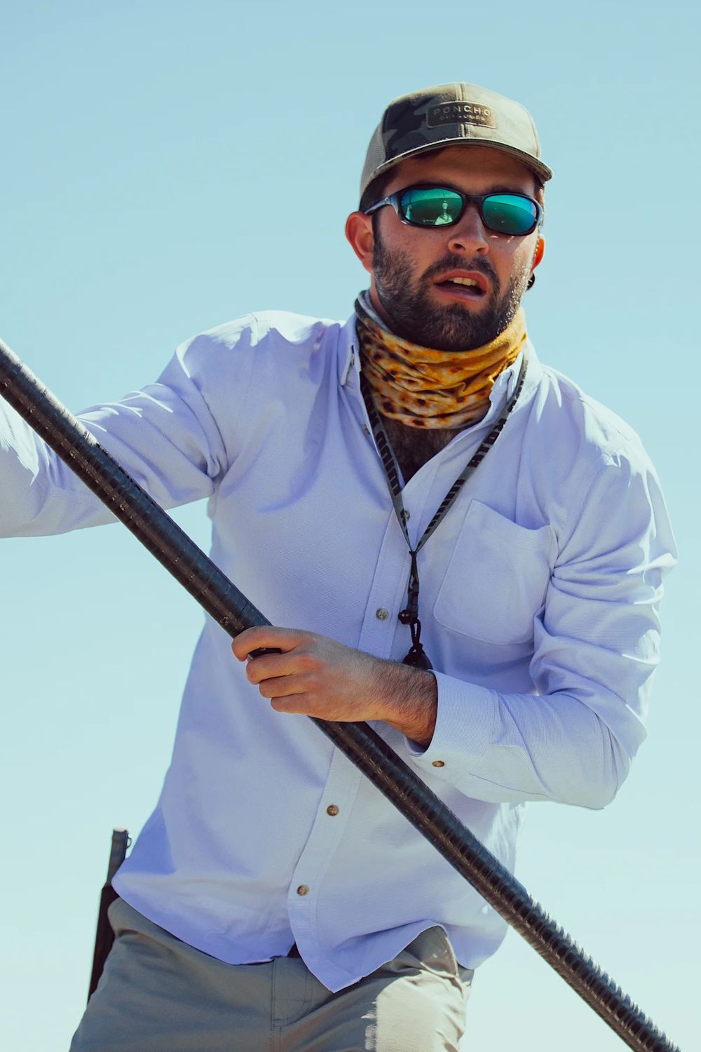 A man wearing a light blue long-sleeve button-up shirt, khaki pants, a brown cap, green-tinted sunglasses, and a yellow patterned neck gaiter, holding a dark metal pole against a clear blue sky.