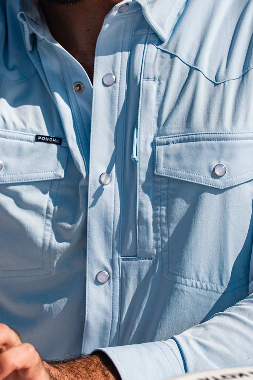 Close-up of a person wearing a light blue button-up shirt with chest flap pockets and silver snap buttons. A small black brand tag on the left chest pocket reads Port & Company.