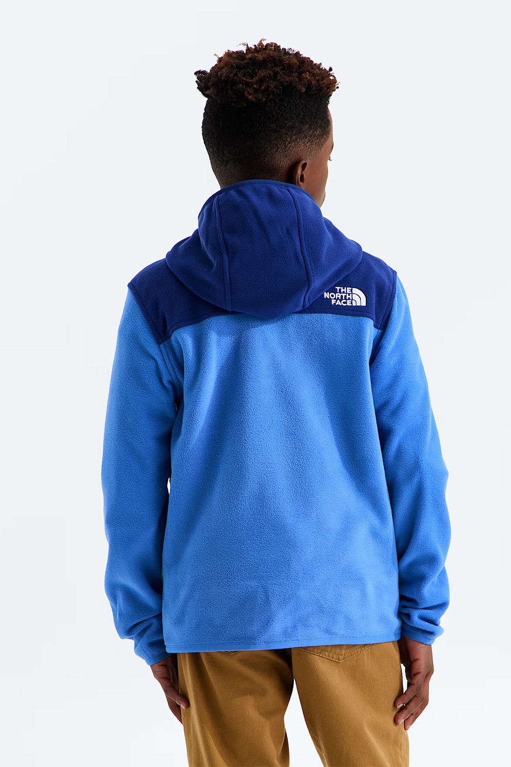 A child viewed from behind wearing a blue and navy hooded fleece jacket with The North Face logo on the back, paired with tan pants, against a plain white studio background.