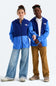 Two models wearing blue and royal blue colorblock zip-up hoodies by The North Face, styled with jeans and brown pants against a light gray studio background.